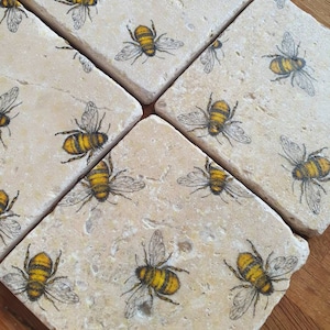May include: Six square stone coasters, each adorned with a detailed illustration of a yellow and black bee. The coasters, with their natural beige stone appearance, are arranged on a wooden surface, offering a rustic aesthetic.