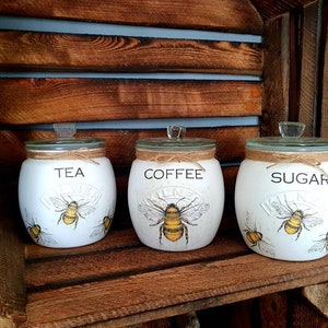 May include: Three white ceramic canisters with bee and floral designs. The canisters are labeled "Tea", "Coffee", and "Sugar".