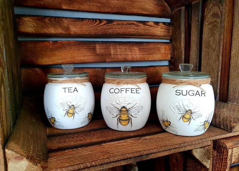 May include: Three white ceramic canisters with bee and floral designs. The canisters are labeled "Tea", "Coffee", and "Sugar".