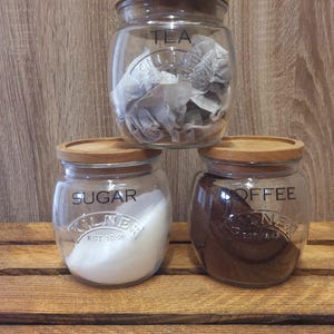 May include: Three glass jars with wooden lids, labelled "TEA", "SUGAR", and "COFFEE". The jars contain tea bags, sugar, and coffee grounds. The jars are stacked on a wooden surface.