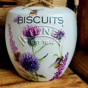 May include: White ceramic biscuit jar with a wooden lid and a twine bow. The jar has a floral design with purple flowers and bees. The text "BISCUITS" is printed on the front of the jar.