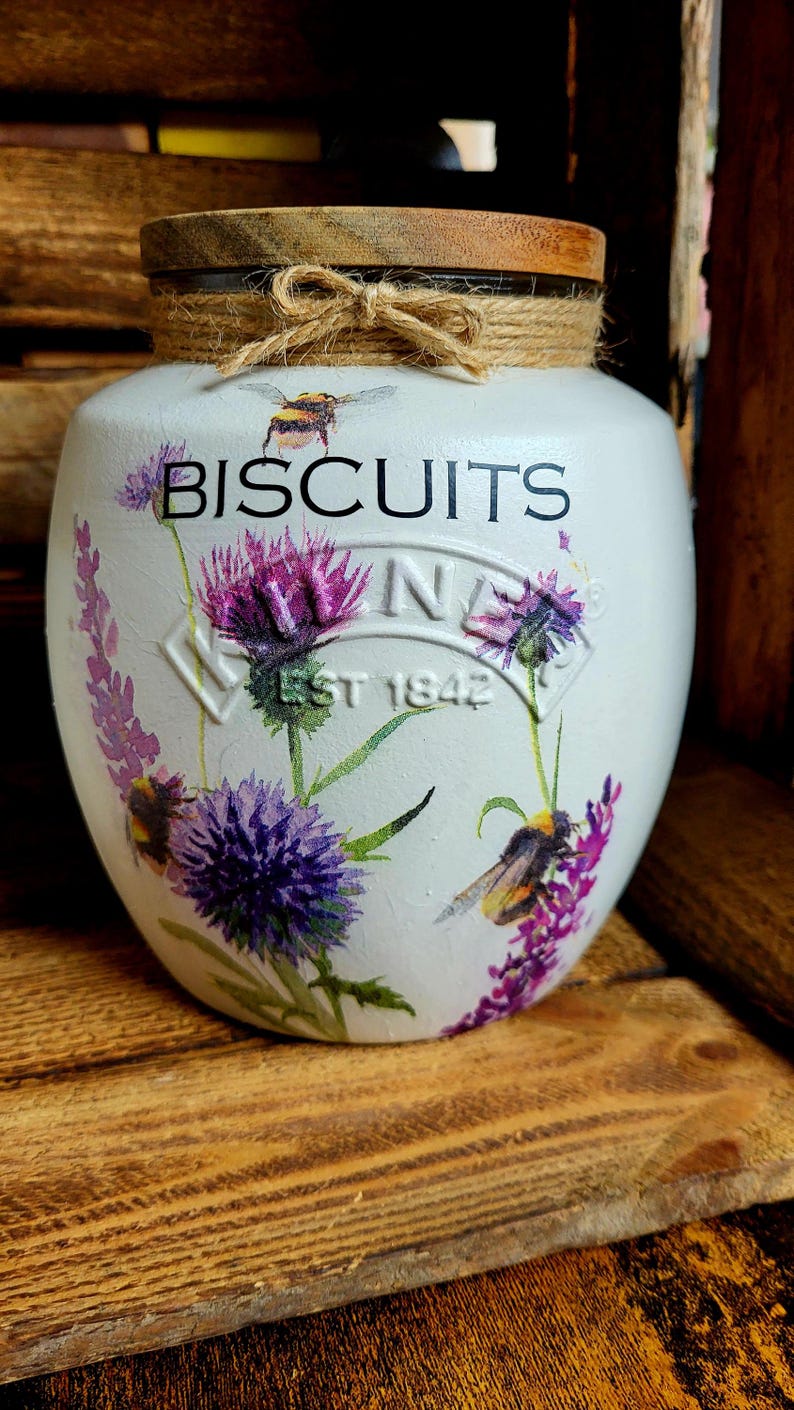May include: White ceramic biscuit jar with a wooden lid and a twine bow. The jar has a floral design with purple flowers and bees. The text "BISCUITS" is printed on the front of the jar.