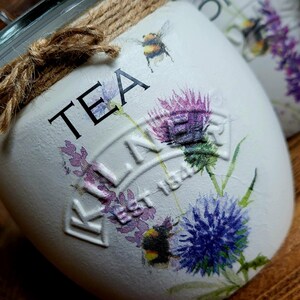 May include: White ceramic tea jar with a brown twine bow and a floral design. The jar has the words "TEA" and "KILNER EST 1842" printed on it.