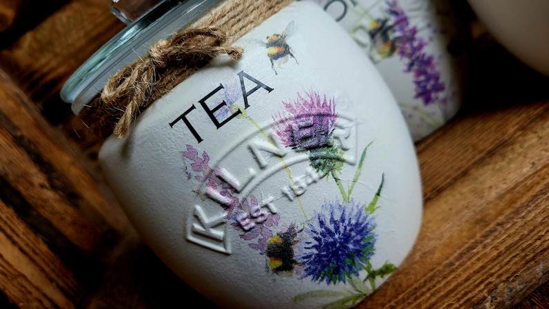 May include: White ceramic tea jar with a brown twine bow and a floral design. The jar has the words "TEA" and "KILNER EST 1842" printed on it.