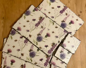 May include: A collection of rectangular and square slate coasters with a floral design. The coasters feature a cream-colored background with purple thistles, lavender, and small bee illustrations. The edges of the coasters are dark gray.