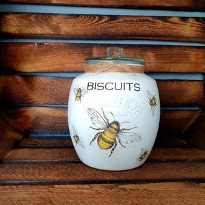 May include: White ceramic biscuit jar with a green lid and a brown twine bow. The jar has a bee and honeycomb design and the text "BISCUITS" printed on the front.