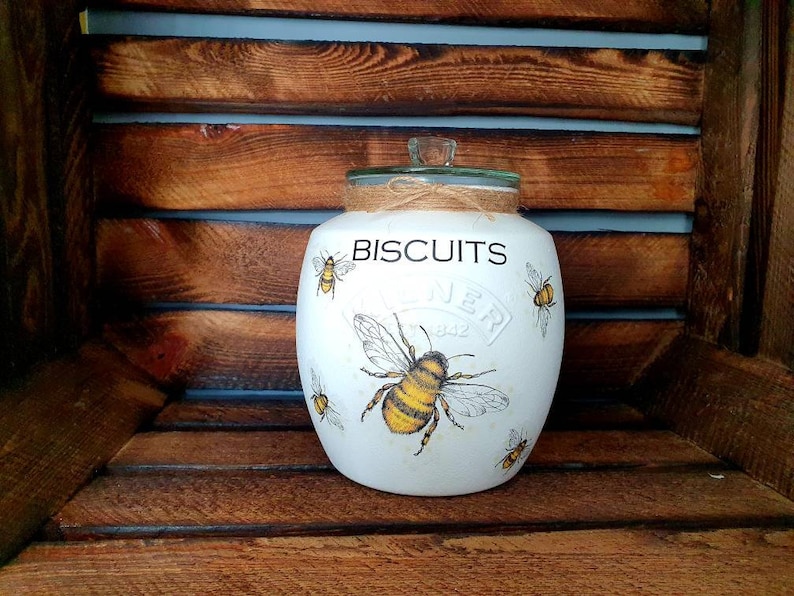 May include: White ceramic biscuit jar with a green lid and a brown twine bow. The jar has a bee and honeycomb design and the text "BISCUITS" printed on the front.
