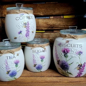 May include: Set of four white ceramic canisters with floral designs and gold accents. The canisters are labeled "Coffee", "Sugar", "Tea", and "Biscuits".