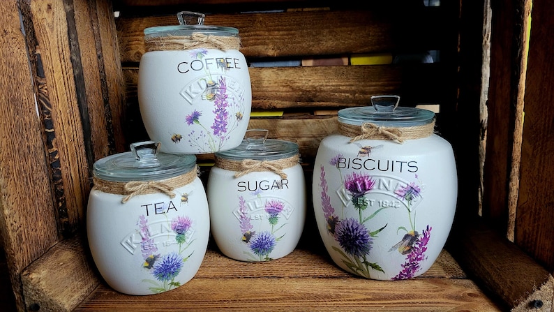 May include: Set of four white ceramic canisters with floral designs and gold accents. The canisters are labeled "Coffee", "Sugar", "Tea", and "Biscuits".