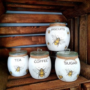 May include: Four white ceramic canisters with bee and floral designs. The canisters are labeled "Tea", "Coffee", "Sugar", and "Biscuits".