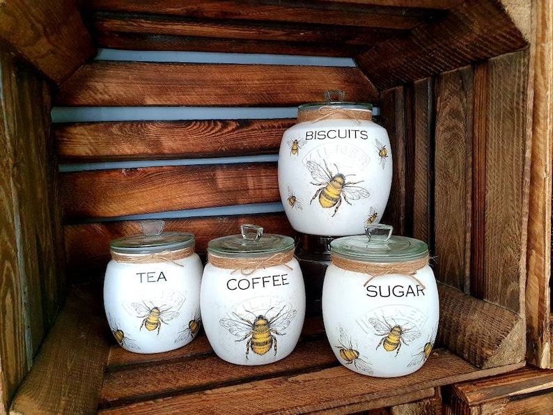 May include: Four white ceramic canisters with bee and floral designs. The canisters are labeled "Tea", "Coffee", "Sugar", and "Biscuits".
