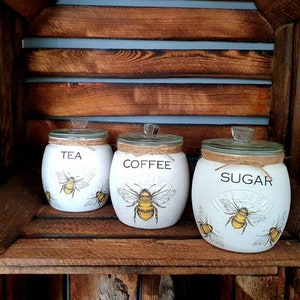 May include: Three white ceramic canisters with bee illustrations and lids. The canisters are labeled "Tea", "Coffee", and "Sugar".