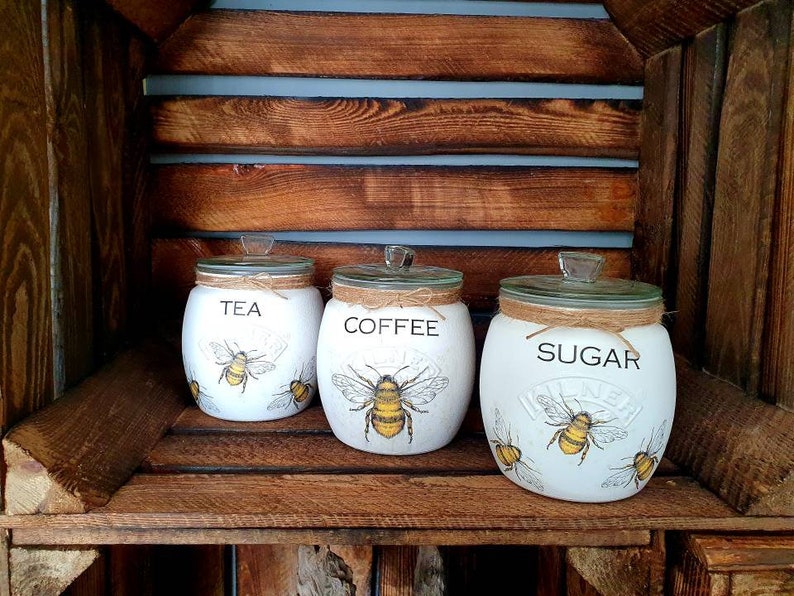 May include: Three white ceramic canisters with bee illustrations and lids. The canisters are labeled "Tea", "Coffee", and "Sugar".