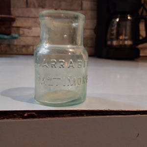 May include: A clear glass bottle with the embossed text "LARRABEE" over "BALTIMORE". The bottle has a cylindrical body and a short neck. The bottle is sitting on a white surface.