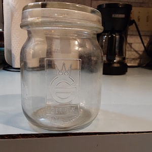May include: A clear glass jar with a wide mouth and a screw-on lid. The jar has a rectangular embossed design with a crown and the word "SEERNX". The jar is sitting on a light blue surface.