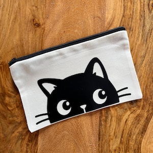 Op de afbeelding: Een rechthoekig, gebroken wit canvas etui met ritssluiting en een zwarte grafische print van een kattengezicht. Het kattenontwerp toont grote ogen, puntige oren en snorharen aan de onderkant van het etui. Het etui heeft een zwarte ritssluiting.