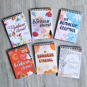Op de afbeelding: Zes spiraalgebonden notitieboekjes met diverse kleurrijke bloemen- en abstracte ontwerpen. Elke kaft bevat de tekst "Un Bonheur éternel" en "ASSEMBLÉE RÉGIONALE 2026" in verschillende lettertypes en lay-outs, waaronder aquarel en lijntekeningen.