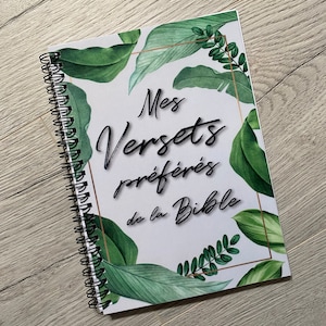 Puede incluir: Una libreta blanca con espiral y un diseño de hojas verdes y doradas. El texto "Mes Versets préférés de la Bible" está impreso en la portada.
