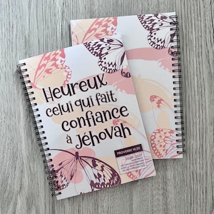 Peut inclure: Deux carnets à reliure spirale avec un fond blanc orné d'illustrations de papillons rose pâle et violet foncé. La couverture affiche le texte "Heureux celui qui fait confiance à Jéhovah" et "Proverbe 16:20 2026-2027 ASSEMBLEE DE CIRCONSCRIPTION AVEC LE RESPONSABLE DE CIRCONSCRIPTION."