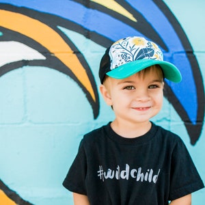 May include: A young child wearing a black t-shirt with the text "#wildchild" printed on it. The child is wearing a turquoise and white patterned trucker hat with a mesh back. The child is standing in front of a wall with a colorful rainbow painted on it.