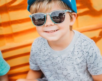 May include: A young boy wearing a blue and white dinosaur patterned baseball cap, clear blue sunglasses, and a gray shirt with a black pattern. He is sitting on a yellow and orange striped surface.