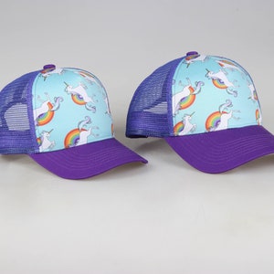 May include: Two purple and blue trucker hats with a light blue background and a white unicorn design with a rainbow.