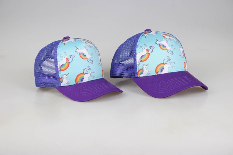 May include: Two purple and blue trucker hats with a light blue background and a white unicorn design with a rainbow.