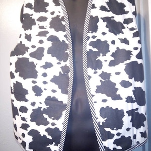 May include: A sleeveless waistcoat featuring a black and white cow print pattern across the front panels. The garment is finished with a black and white diagonal striped trim along the edges of the neckline, front opening, and the bottom hemline.