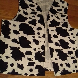 May include: A sleeveless vest featuring a cow print pattern. The vest is white with large black blotches, and has a V-neck. The garment is open at the front and appears to be made of fabric.