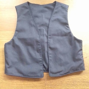 Pode incluir: Um colete cinza liso com decote em V. A peça sem mangas é feita de um tecido liso e tem um design simples e clássico. Está deitado em uma superfície de madeira. Uma peça de roupa versátil.