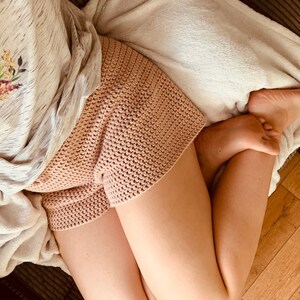 May include: A close-up of a person wearing a pair of pink crocheted shorts. The shorts are made with a simple stitch pattern and have a relaxed fit.