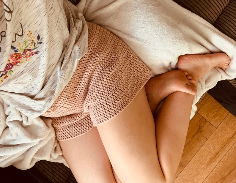May include: A close-up of a person wearing a pair of pink crocheted shorts. The shorts are made with a simple stitch pattern and have a relaxed fit.