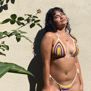 May include: A woman wearing a colorful crocheted bikini top and matching bottom. The bikini is made with a variety of colors, including yellow, orange, purple, and blue. The woman is standing in front of a white wall with a green plant in the foreground.