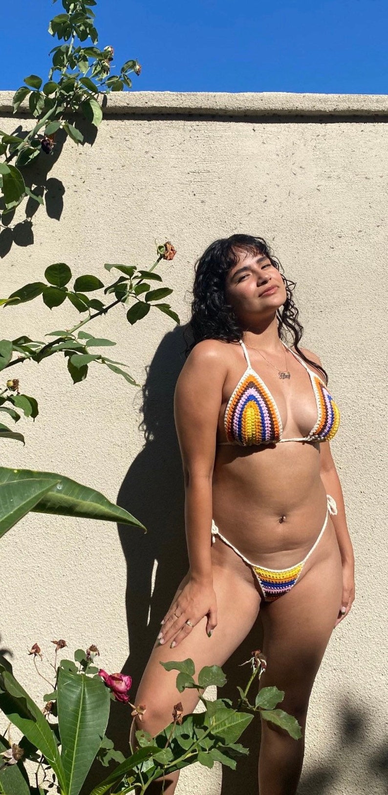 May include: A woman wearing a colorful crocheted bikini top and matching bottom. The bikini is made with a variety of colors, including yellow, orange, purple, and blue. The woman is standing in front of a white wall with a green plant in the foreground.