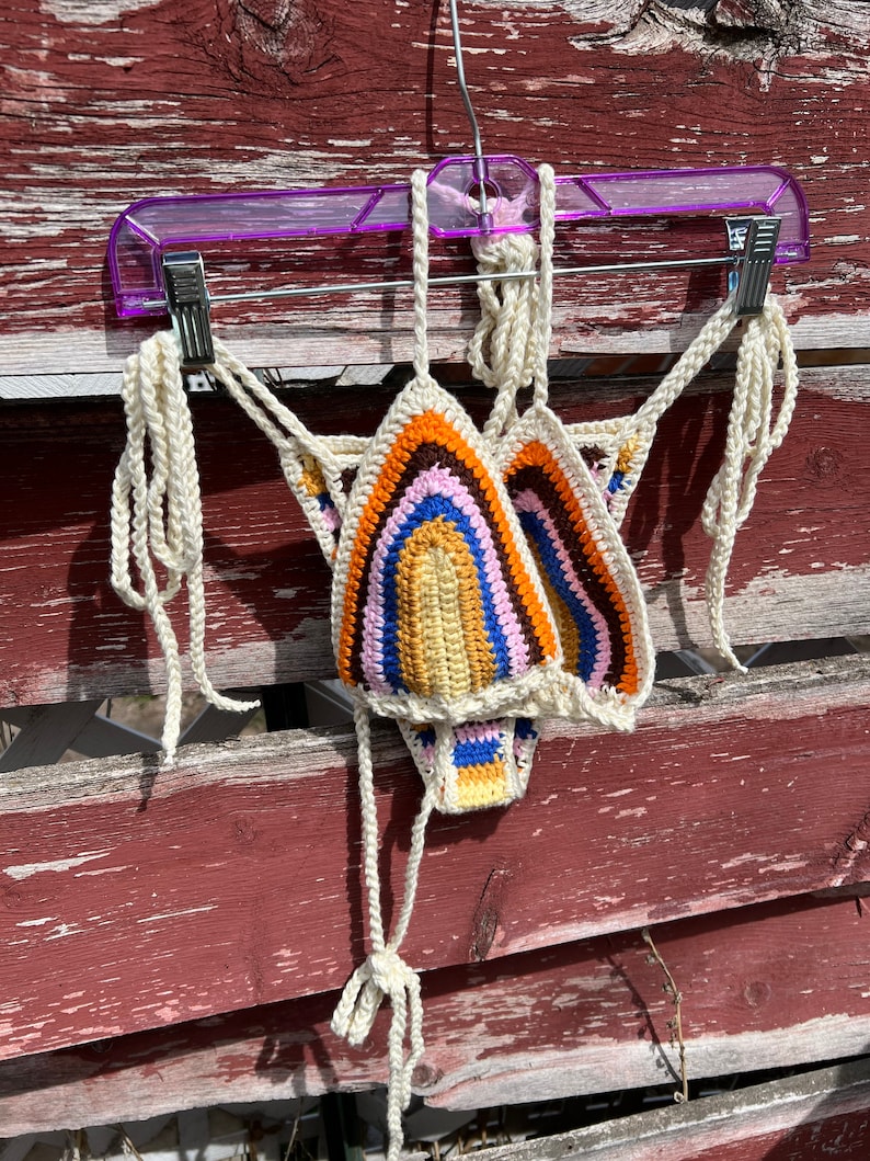 May include: A rainbow crochet bikini top with white straps hanging on a purple hanger. The top is made of a soft, colorful yarn and has a unique, bohemian style.