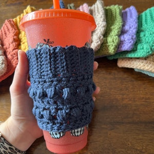 May include: A close-up of a crocheted cup cozy in navy blue, with a red and white patterned reusable cup with a straw. The cozy is made with a textured stitch and is designed to fit a standard-sized cup.