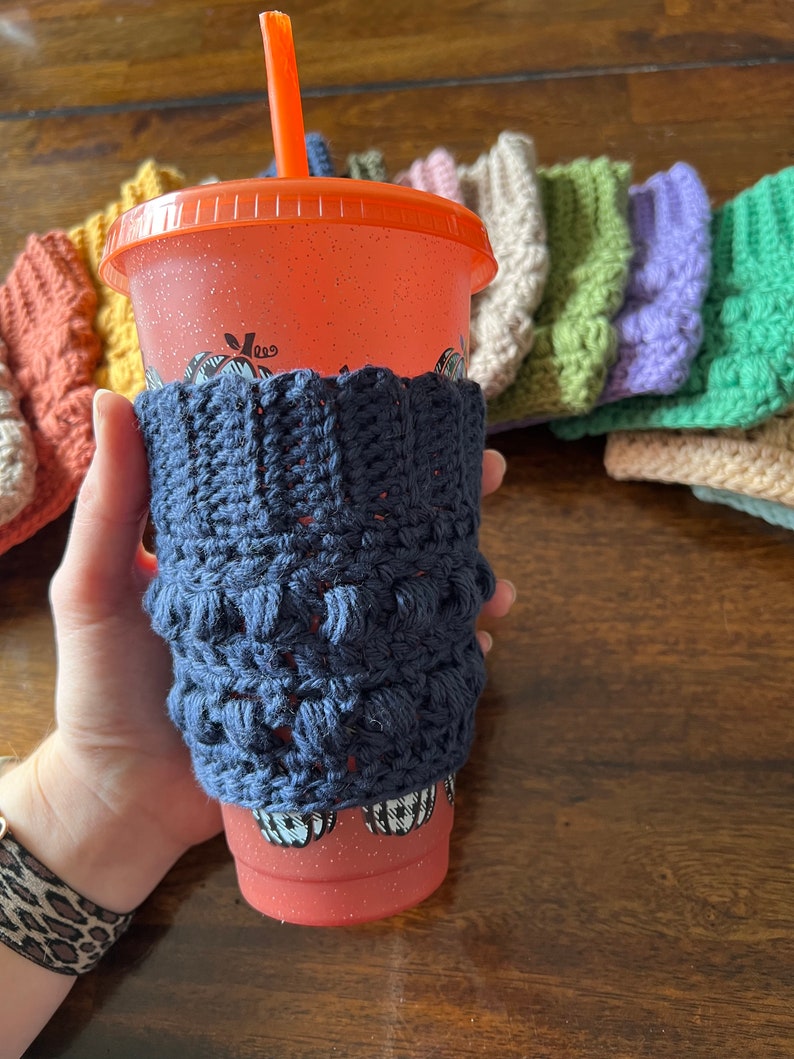 May include: A close-up of a crocheted cup cozy in navy blue, with a red and white patterned reusable cup with a straw. The cozy is made with a textured stitch and is designed to fit a standard-sized cup.