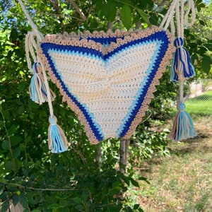 May include: A crocheted heart-shaped bag with blue, beige, and white stripes. The bag has tassels on the ends of the straps.