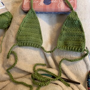 Puede incluir: Un top de bikini de crochet verde con lazos largos. El top est&aacute; colocado sobre una superficie blanca y peluda.