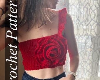 Puede incluir: Un top de crochet rojo con una gran rosa tridimensional en la parte delantera. El top es sin mangas y tiene un escote con volantes. La modelo lleva pantalones vaqueros azules.