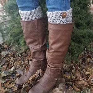 May include: Brown leather boots with a pair of beige crocheted boot cuffs with a button on the cuff. The boots and cuffs are worn by a person standing on a bed of brown leaves.