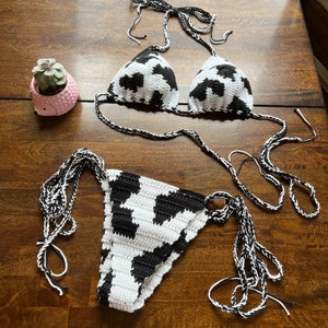 May include: A two-piece crochet bikini set featuring a black and white cow print pattern. The top consists of two triangular cups with braided tie straps, and the bottom is a matching piece with side ties. A small pink succulent planter is nearby.