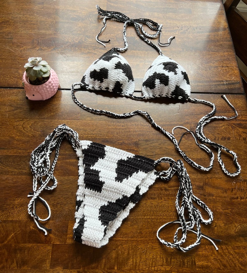 May include: A two-piece crochet bikini set featuring a black and white cow print pattern. The top consists of two triangular cups with braided tie straps, and the bottom is a matching piece with side ties. A small pink succulent planter is nearby.