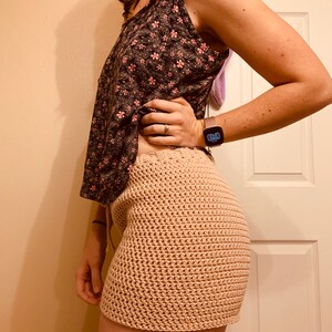 May include: A woman wearing a floral print tank top and a crocheted skirt. The skirt is a light beige color and has a loose, flowy fit.
