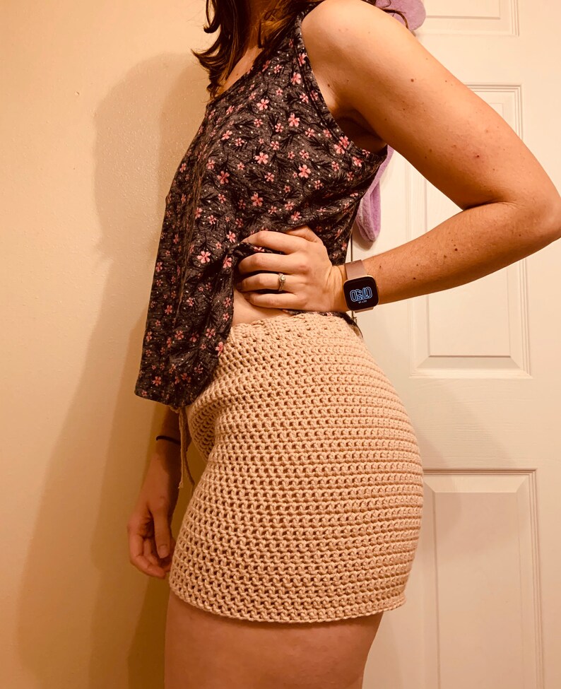 May include: A woman wearing a floral print tank top and a crocheted skirt. The skirt is a light beige color and has a loose, flowy fit.