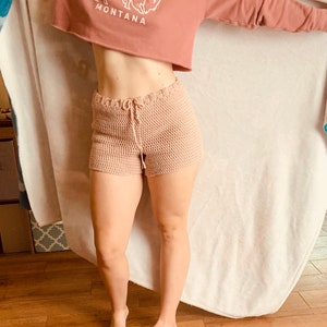 May include: A woman wearing a pink cropped sweatshirt with a white buffalo graphic and the word "MONTANA" printed on it. She is also wearing a pair of light pink crochet shorts.