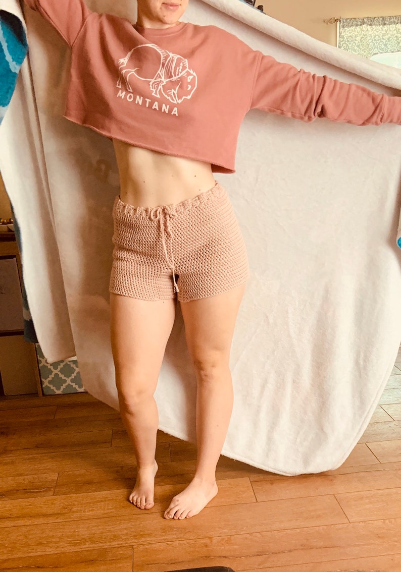 May include: A woman wearing a pink cropped sweatshirt with a white buffalo graphic and the word "MONTANA" printed on it. She is also wearing a pair of light pink crochet shorts.