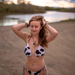 May include: A two-piece crochet bikini set featuring a white base with black cow-print patterns. The top is a triangle style with thin straps, and the bottom features side-tie closures with matching black and white braided strings.