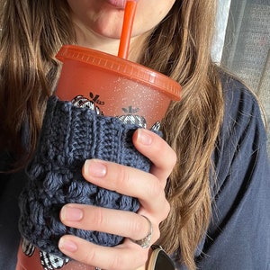 May include: A close-up of a person's hand holding a reusable orange and white tumbler with a black and white pumpkin design. The tumbler has a knitted gray cozy and a red straw.