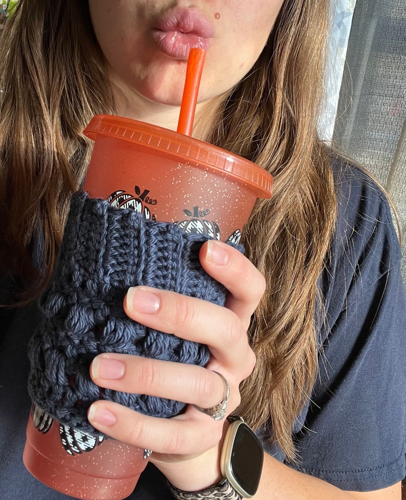 May include: A close-up of a person's hand holding a reusable orange and white tumbler with a black and white pumpkin design. The tumbler has a knitted gray cozy and a red straw.